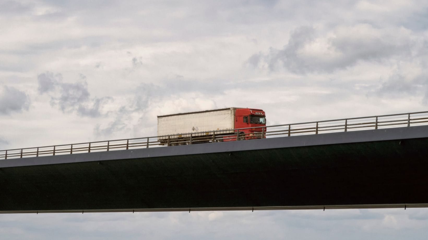 Red lorry crossing a long concrete bridge under a cloudy sky