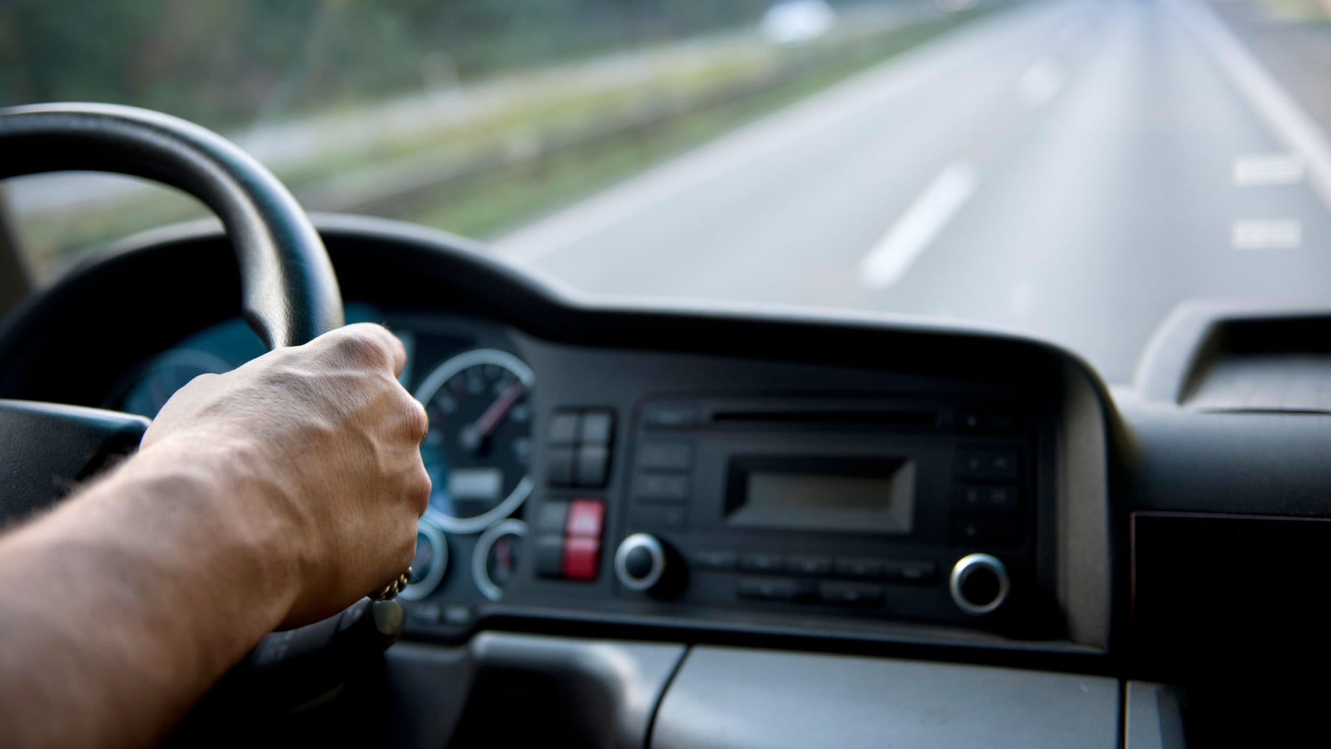 Hand van een vrachtwagenchauffeur op het stuur, met het dashboard en een lege snelweg voor zich