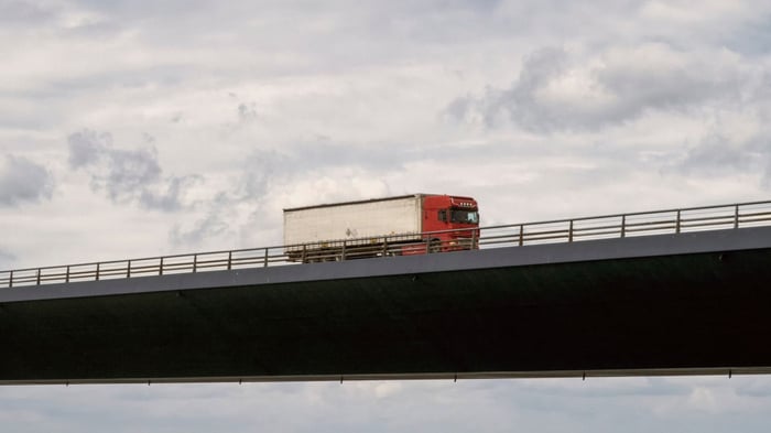 Red lorry crossing a long concrete bridge under a cloudy sky